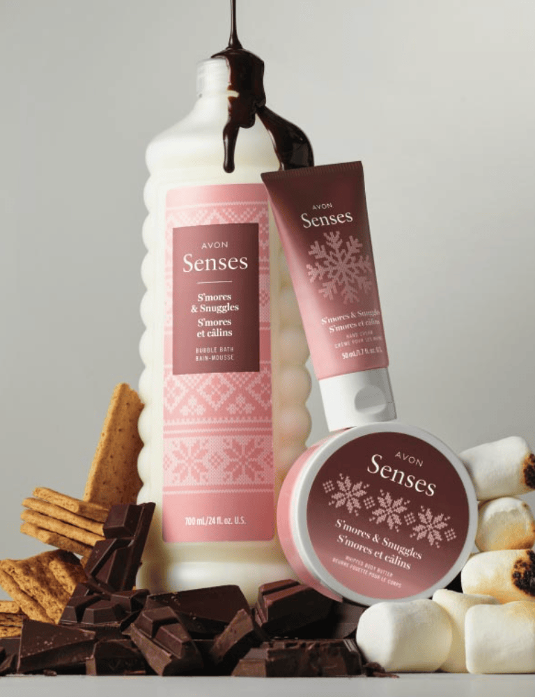A collection of Avon Senses bath products featuring 'S'mores & Snuggles' bubble bath, body cream, and body butter, displayed with chocolate, graham crackers, and marshmallows on a neutral background.